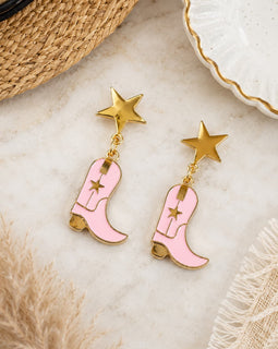 Pink cowboy boot earrings with gold stars on a textured surface.