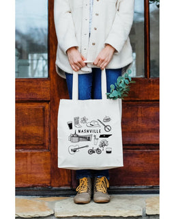 Person holding a tote bag with 'Nashville' design in front of a wooden door.