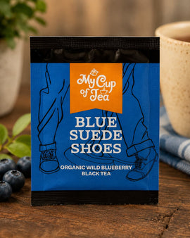 Tea package labeled 'Blue Suede Shoes' by My Cup of Tea on a wooden surface with blueberries and a cup.