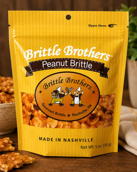 Yellow package of Brittle Brothers Peanut Brittle on a wooden surface with a bowl of brittle and a plant in the background.