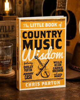 Book titled 'The Little Book of Country Music Wisdom' by Chris Parton on a wooden surface with a guitar and glass in the background.