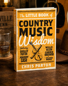 Book titled 'The Little Book of Country Music Wisdom' by Chris Parton on a wooden surface with a guitar and glass in the background.