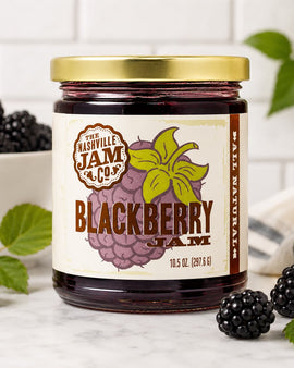 Jar of Nashville Jam Co. Blackberry Jam with blackberries and leaves on a marble surface.