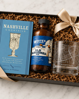 Gift set with 'Nashville Cocktails' book, bottle of 'White's Elixirs Old Fashioned', and glass in a box on a marble surface.
