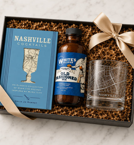Gift set with 'Nashville Cocktails' book, bottle of 'White's Elixirs Old Fashioned', and glass in a box on a marble surface.