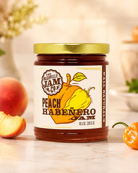 Jar of Peach Habanero Jam from Nashville Jam Co. with peaches and habanero peppers on a wooden surface.