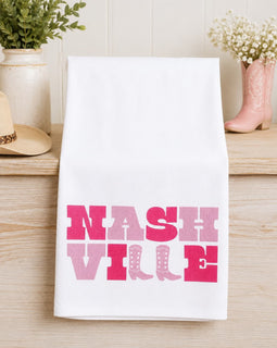 White towel with 'NASH VILLE' printed in pink on a wooden surface with decorative items.