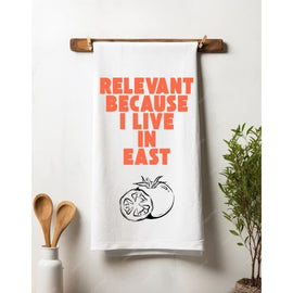 White cotton tea towel with black screen-printed text reading “Relevant because I live in East Nashville,” styled on a rustic wood counter.