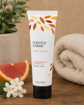 Thistle Farms body lotion tube with grapefruit and jasmine flowers on a wooden surface.