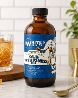 Bottle of White's Elixirs Old Fashioned Mix on a kitchen counter with a glass of cocktail and bar tools.