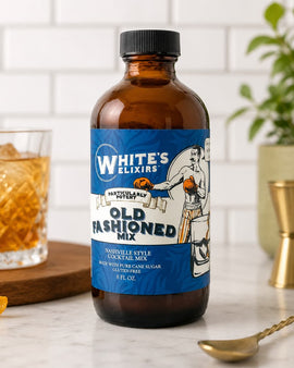 Bottle of White's Elixirs Old Fashioned Mix on a kitchen counter with a glass of cocktail and bar tools.